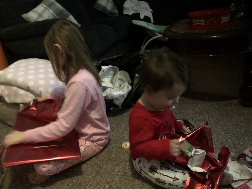 Lottie and Mia sat back to back opening presents wrapped in red paper