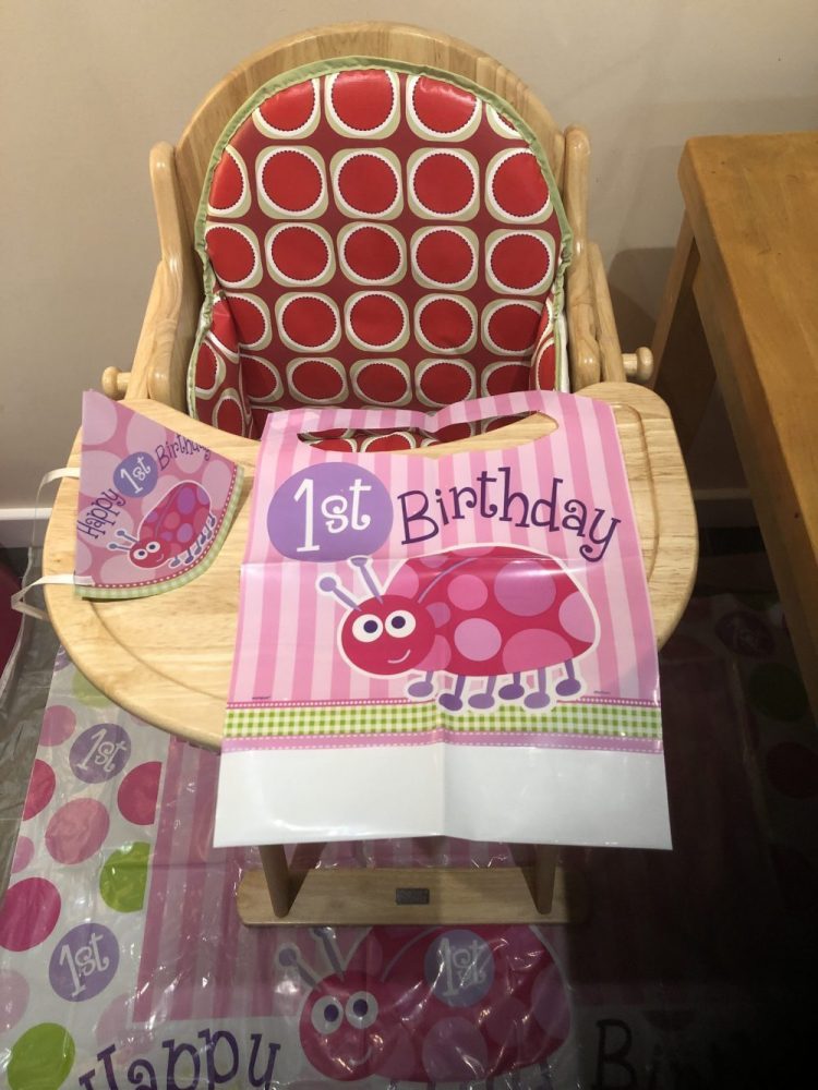 Wooden High Chair with red patterned insert. Decorated with 1st birthday banner, hat and bib with a pink ladybird on it