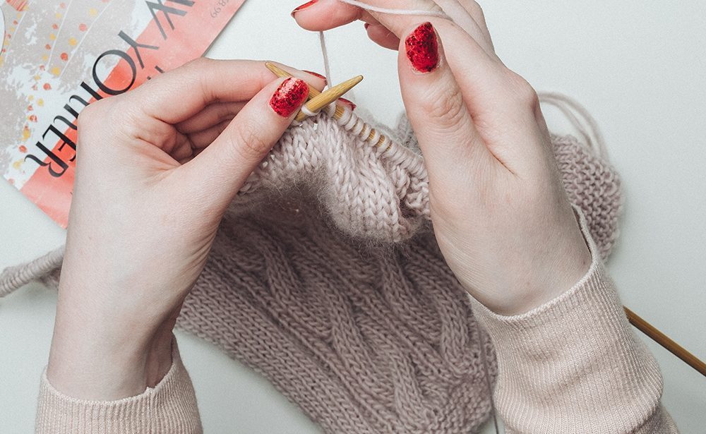 2 hands with red painted nails holding knitting needles, knitting with beige wool