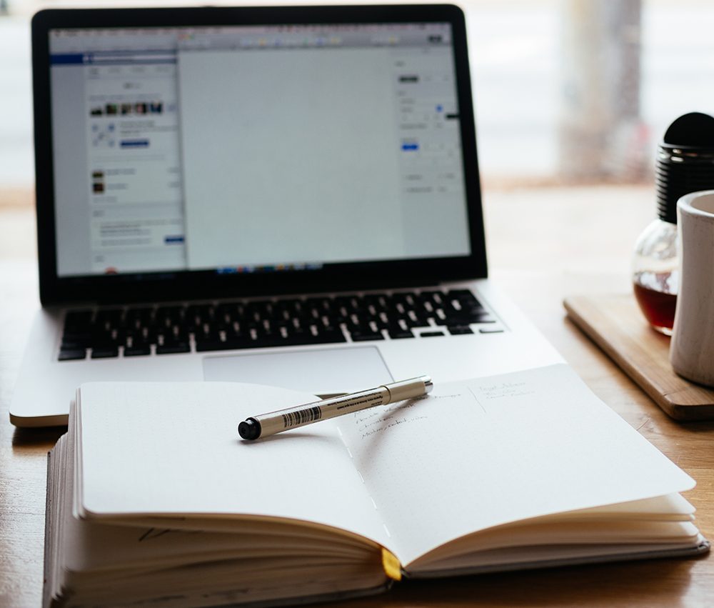 laptop open in the background, silver pen resting on an open notebook