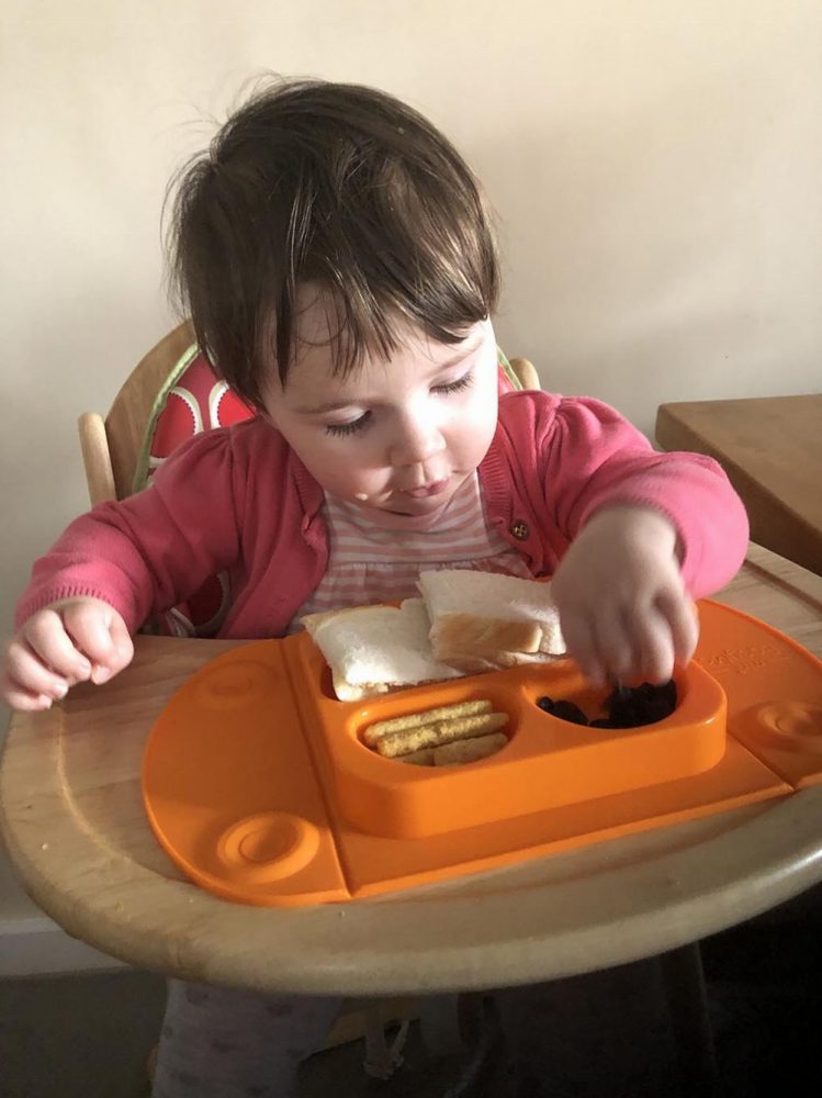 Lottie eating her lunch from an orange Mini EasyMat. Suctioned to a wooden high chair tray. Top Baby Product - EasyTots